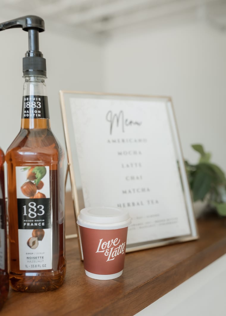 A printed drinks menu in a frame beside a syrup bottle and a branded cup
